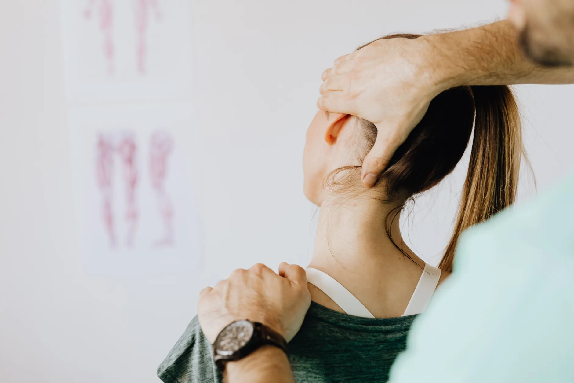A practitioner examining the neck and shoulder of a seated patient, with a spinal anatomy chart on the wall behind
